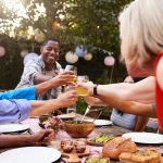 Friends toast with glasses over a table of food during an outdoor garden party decorated with paper lanterns.